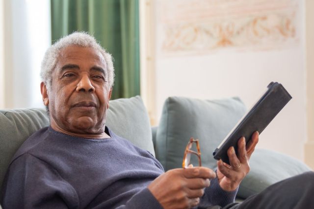 Older man sitting on a sofa at home holding a tablet and reading glasses