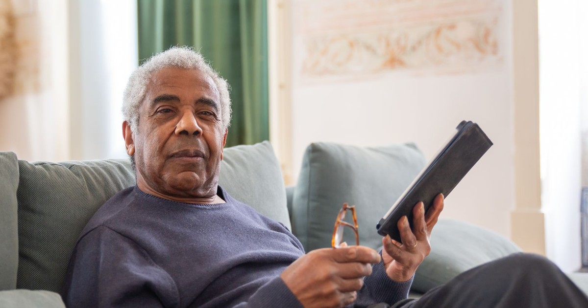 Older man sitting on a sofa at home holding a tablet and reading glasses
